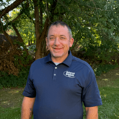 Portrait of a smiling man wearing a navy blue polo shirt with company logo, standing outdoors against green foliage