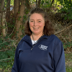 Woman with long brown hair smiling at camera wearing a dark blue polo shirt with embroidered logo, outdoors with trees in background