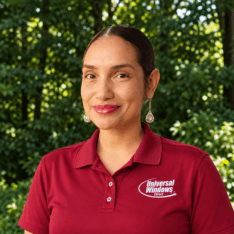 Woman with dark hair wearing a maroon polo shirt with logo, smiling at camera with green foliage background