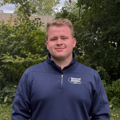 Man wearing dark blue quarter-zip pullover with company logo, smiling at camera outdoors with green foliage background